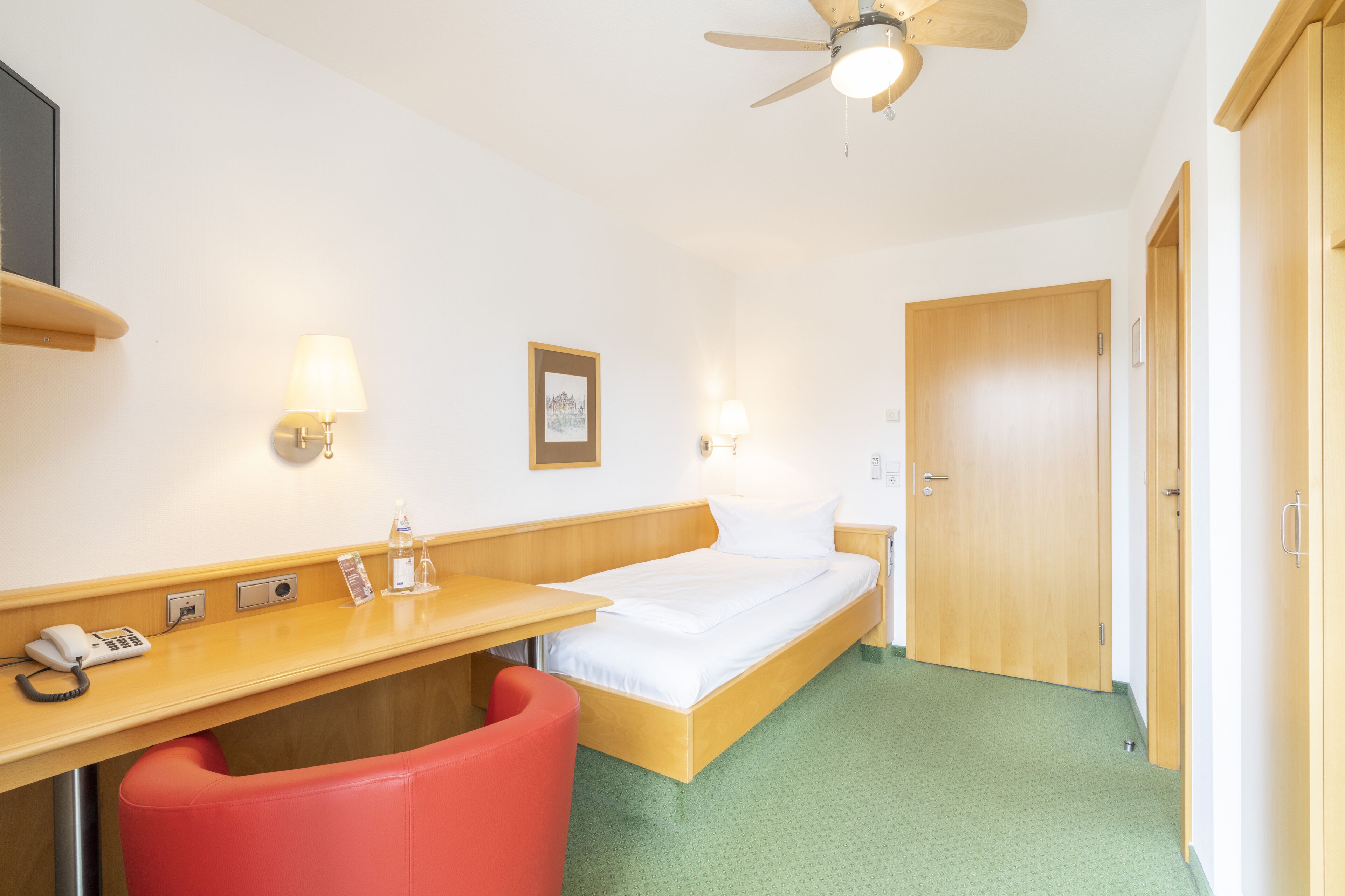A hotel room with a bed, a desk with a phone, a red chair, a ceiling fan, and a door.
