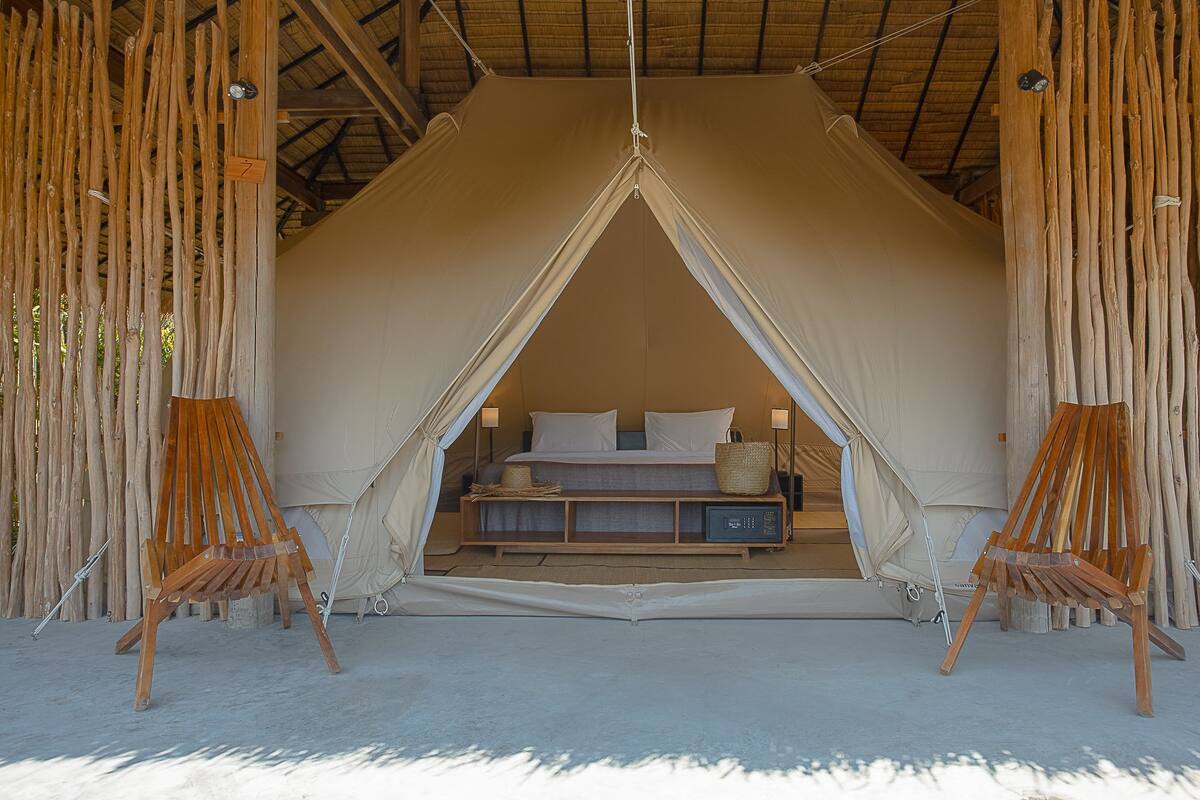 A tented accommodation with a bed, bedside table, and two chairs.