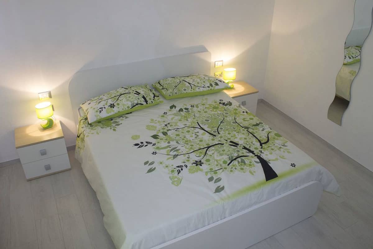Un lit bien fait, recouvert d’une courtepointe ornée d’arbres et de fleurs, deux lampes de chevet, une petite table de chevet et un miroir accroché au mur.