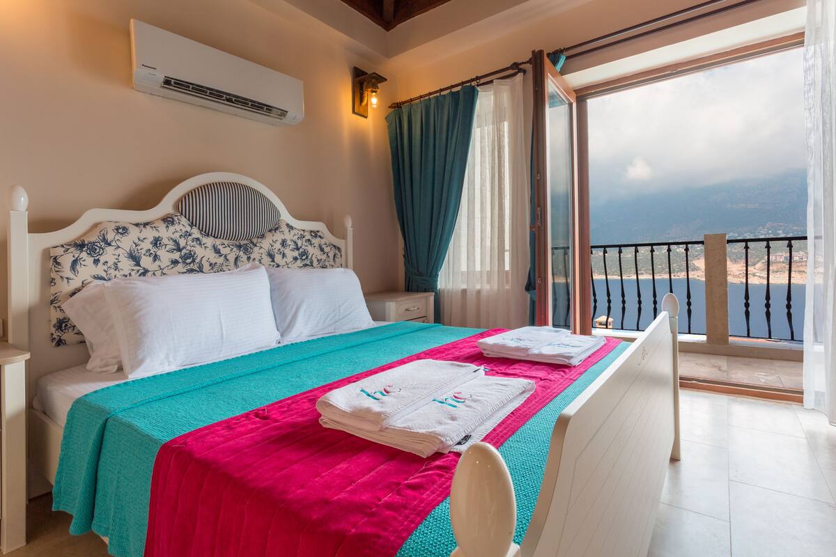 Een slaapkamer met een bed, een nachtkastje, een raam met gordijnen en uitzicht op zee.