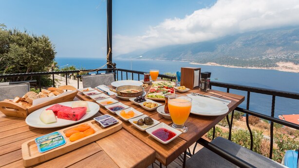 Ein Holztisch mit Frühstücksleckereien, darunter Brot, Obst und ein Glas Orangensaft, mit Blick auf eine malerische Küstenlandschaft.