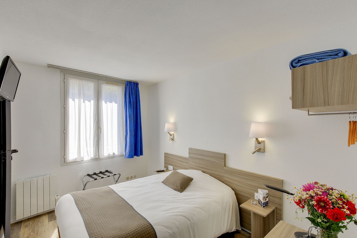 Ein Hotelzimmer mit Bett, Nachttisch, Lampe und einem Fenster mit Vorhängen.