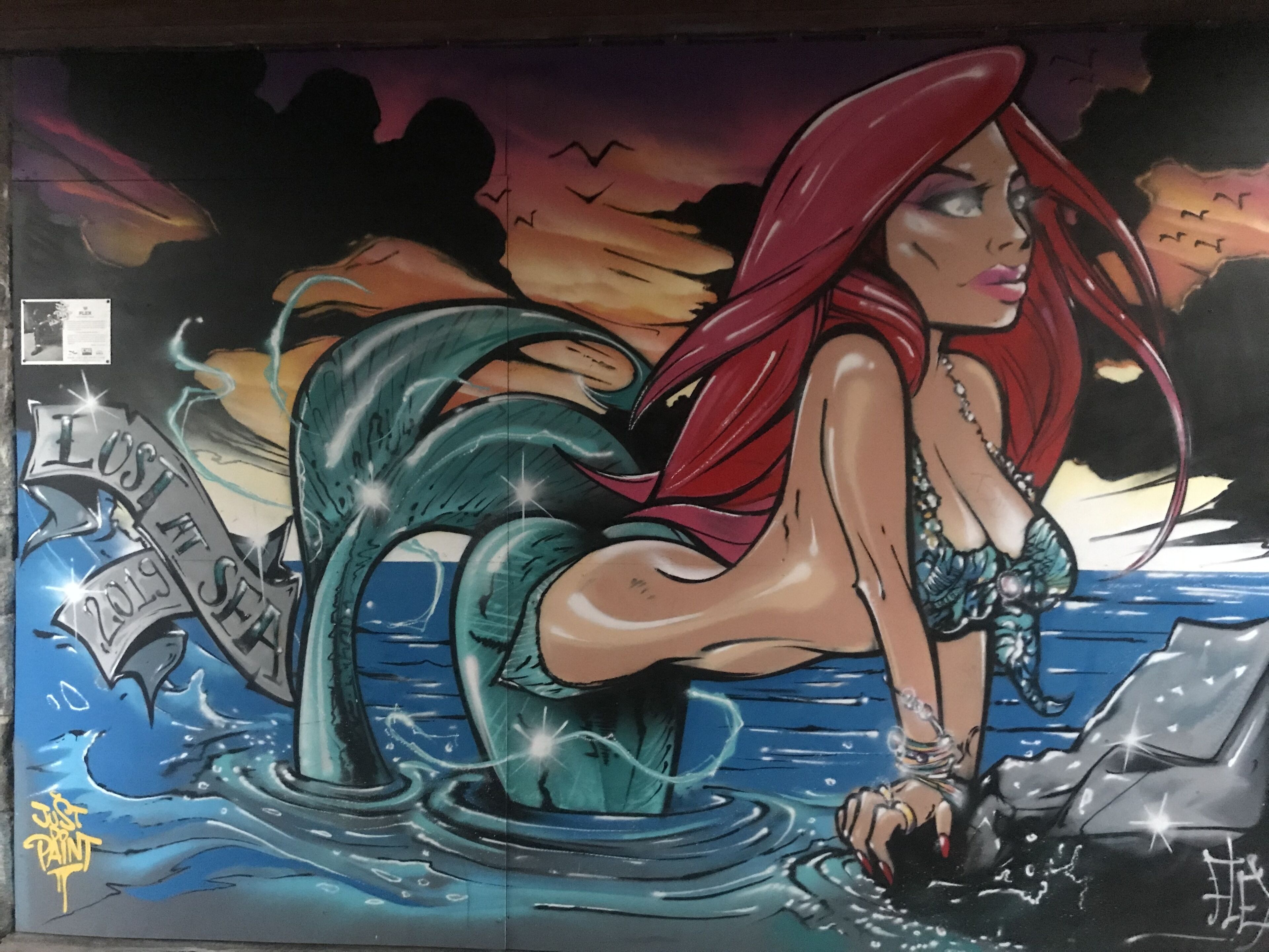 Um mural mostrando uma sereia de cauda vermelha e capa vermelha, sentada na água, com um navio ao fundo.
