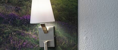 A wall-mounted outdoor lamp with a rectangular shade, set against a backdrop of lush greenery and purple flowers.