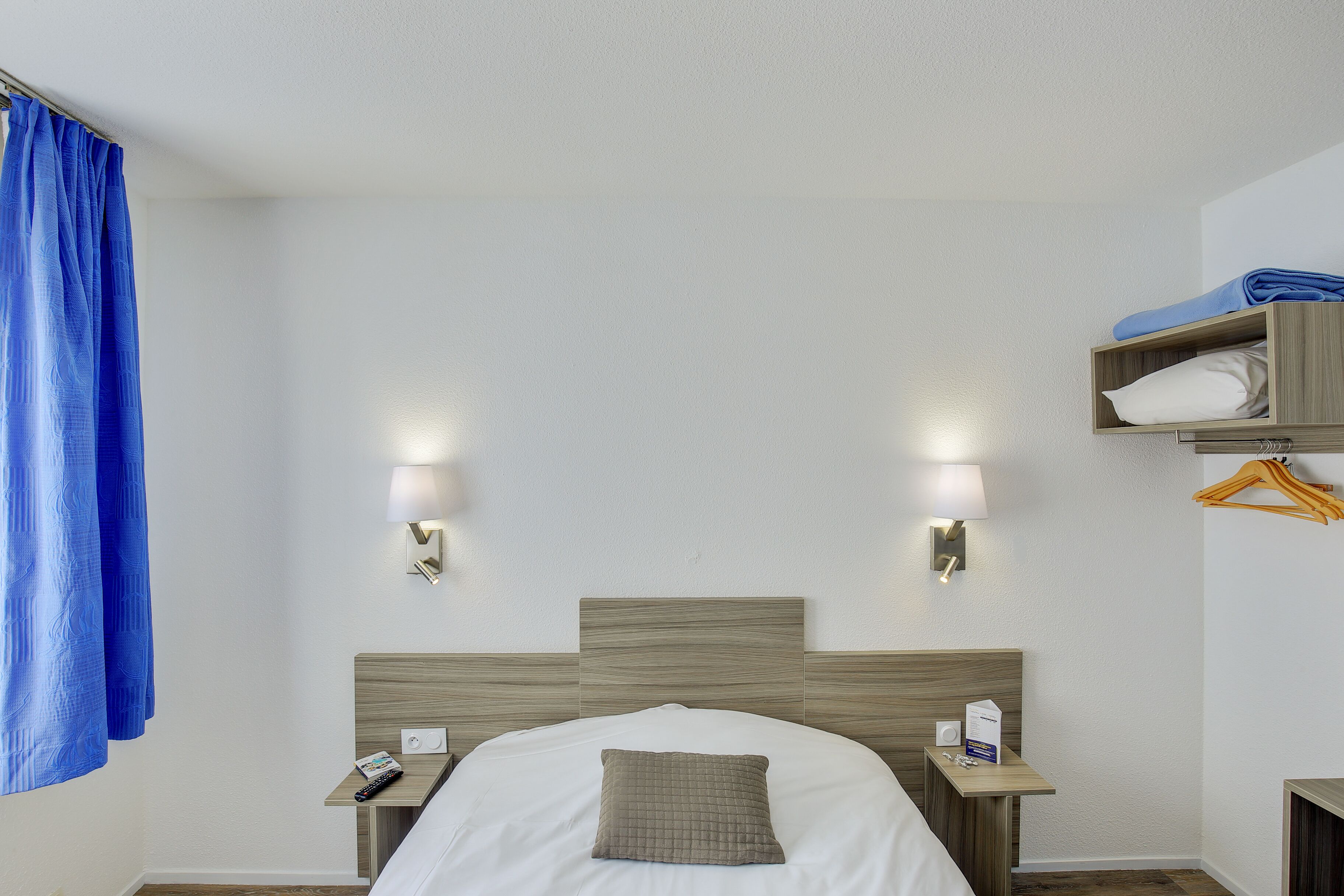 A hotel room with a bed, bedside tables, a lamp, a hanging blue curtain, and a wooden shelf with folded clothes.