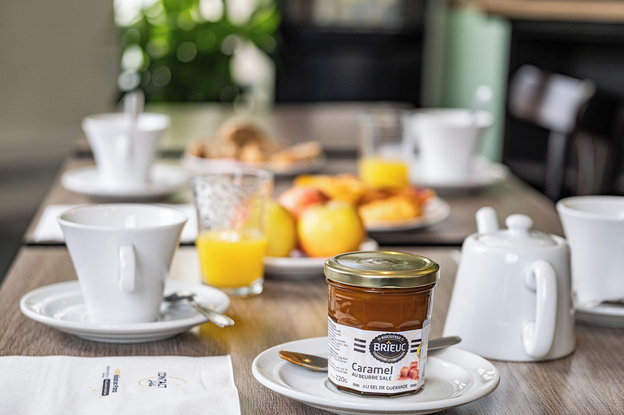A breakfast setting with a jar of Brieuc caramel honey, white cups, a glass of orange juice, and a plate of fruit.