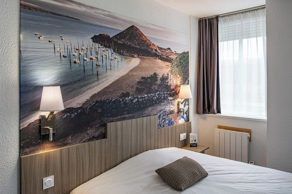 Ein Hotelzimmer mit einer großen Wandmalerei einer Strandansicht, einem Bett mit Kissen, zwei Nachttischlampen und einem Fenster mit Vorhängen.