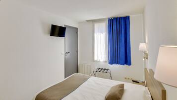 A hotel room with a bed, a TV mounted on the wall, a window with blue curtains, a radiator, and a bedside table with a lamp.
