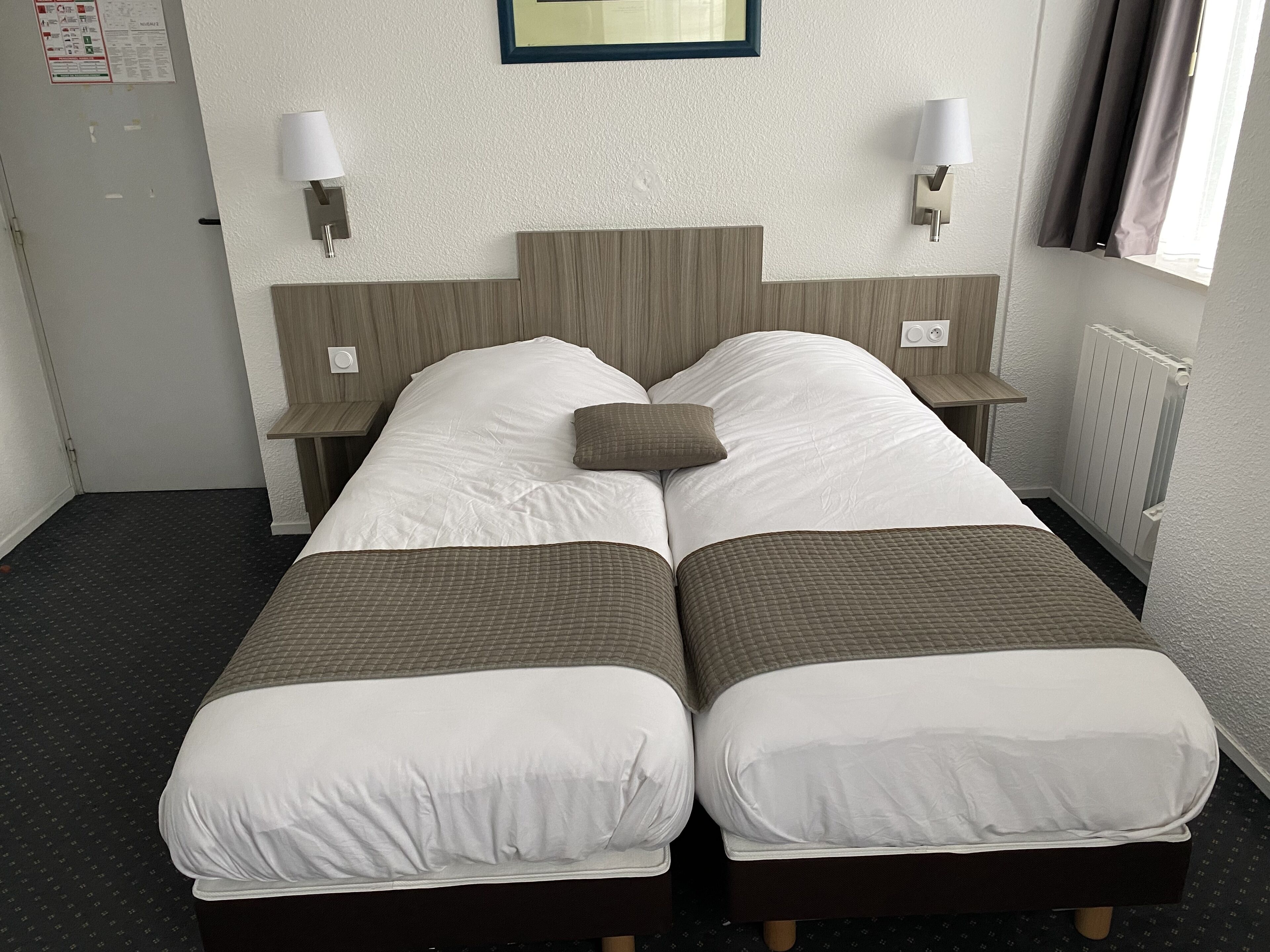 Una cama doble con dos almohadas, una mesita de noche, una lámpara de pared y un ventanal con cortinas.