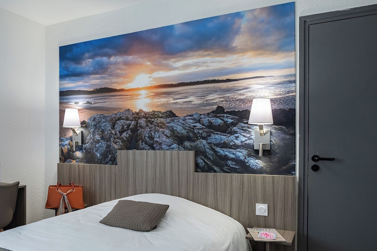 Ein Hotelzimmer mit großem Wandbild, einem Bett mit Kissen, einem Nachttisch, einer Lampe und einer Handtasche an der Wand.