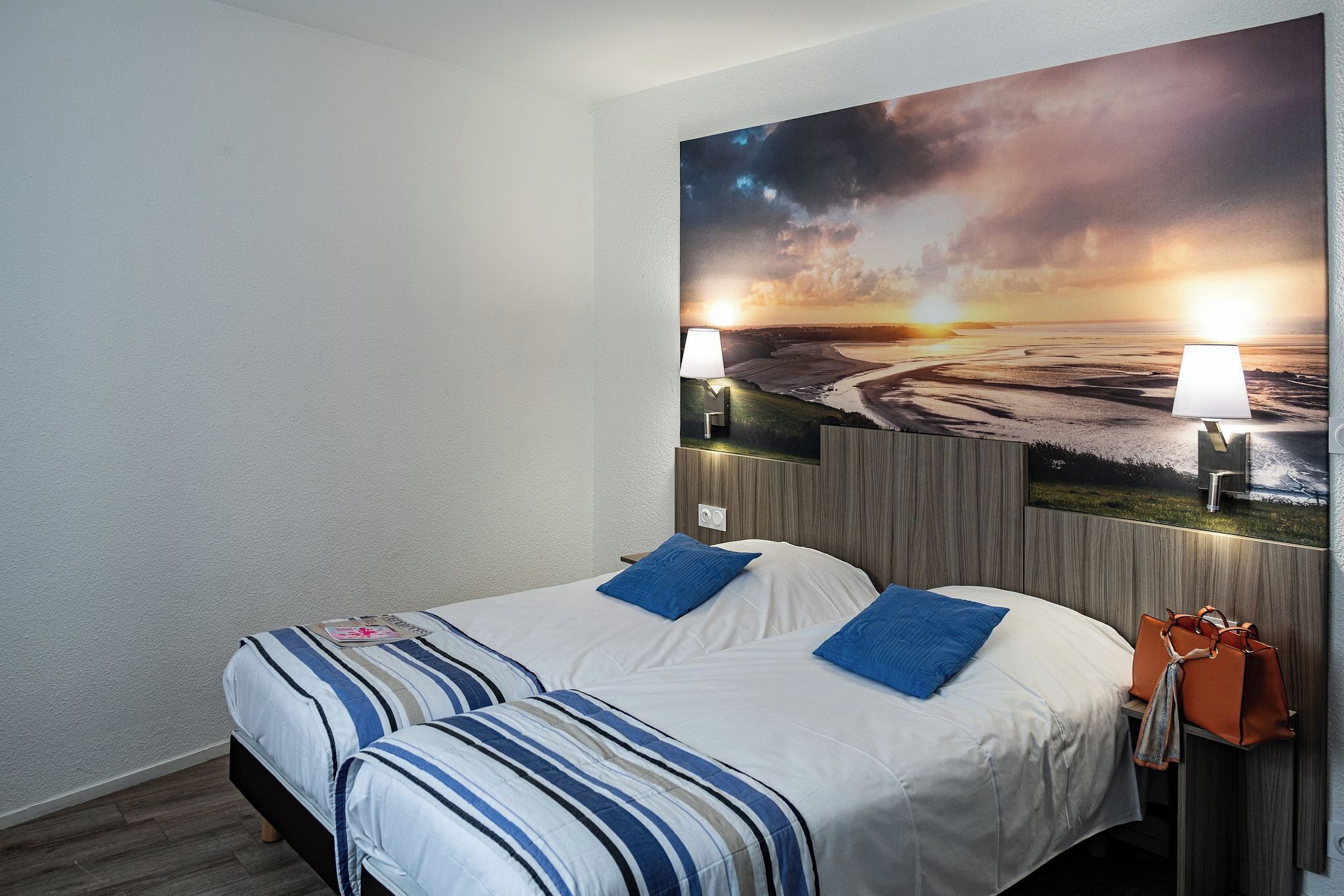 Une chambre d’hôtel avec un lit, deux lampes, un tableau représentant un coucher de soleil et un sac à main posé sur la table de chevet.