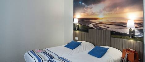 Une chambre d’hôtel avec un lit, deux lampes, un tableau représentant un coucher de soleil et un sac à main posé sur la table de chevet.