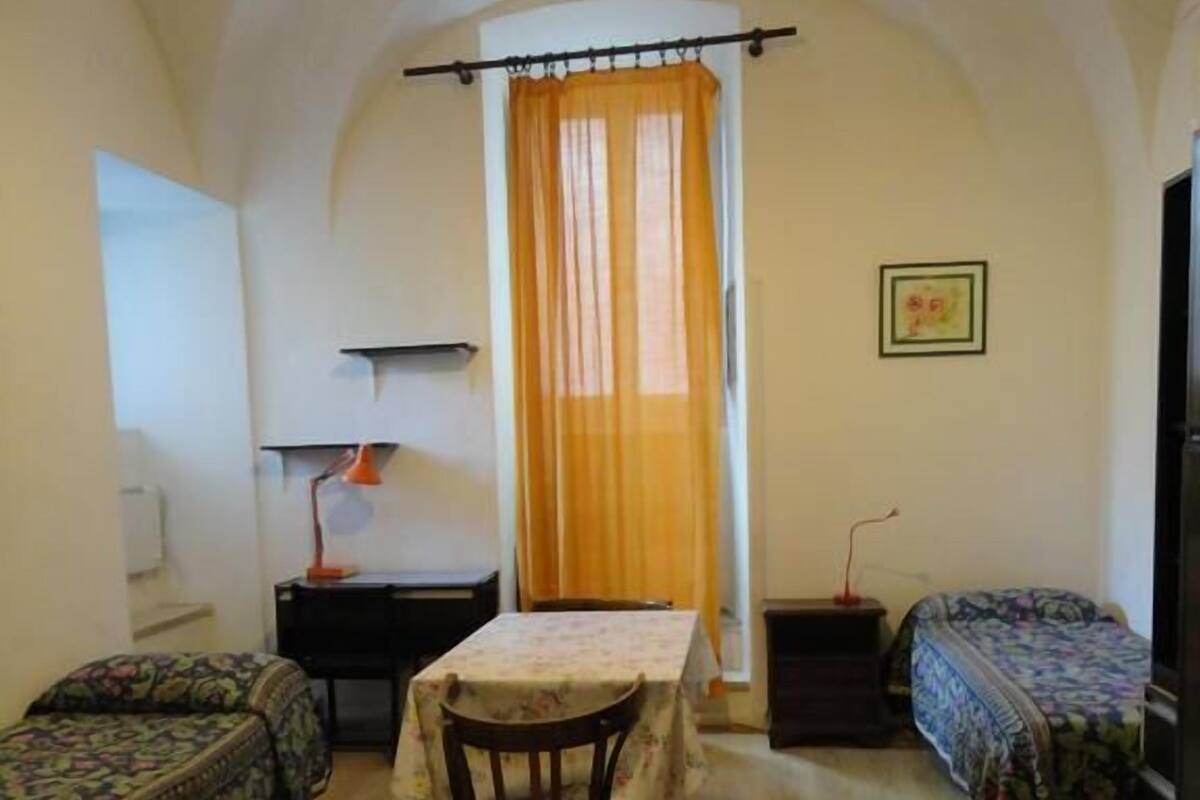 Una habitación pequeña y acogedora con suelo de madera, una mesa con mantel floral, dos camas con ropa de cama estampada, un escritorio con lámpara y una ventana con cortinas naranjas.