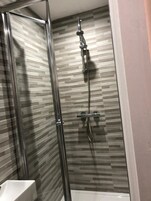Una ducha con cerramiento de vidrio, pared con azulejos y una bañera blanca.