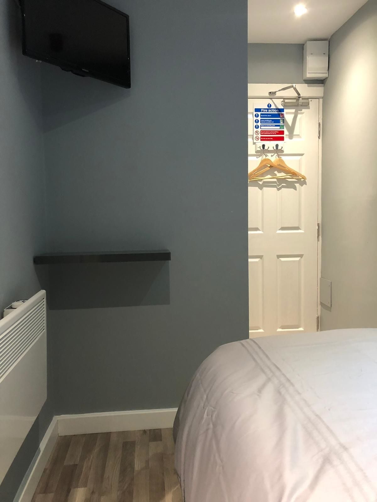 A bedroom with a bed, a mounted TV, a wall-mounted shelf, and a door with a fire action sign.
