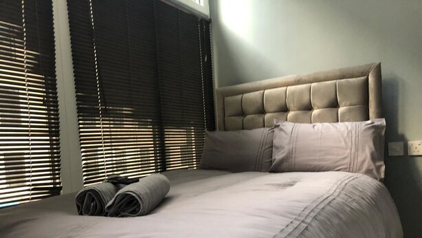 A neatly made bed with a tufted headboard, a pair of rolled towels, and a window with blinds. - Brownlows Inn (Liverpool)
