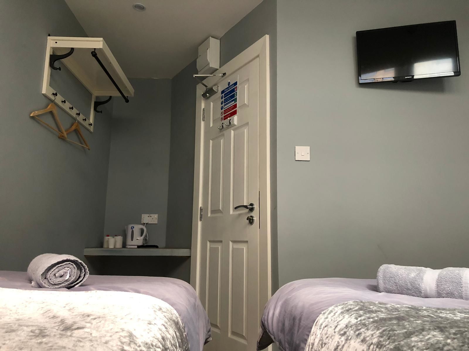 A bedroom with two beds, a TV mounted on the wall, a door with a fire safety sign, and a small shelf with a kettle.