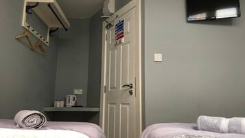 A bedroom with two beds, a TV mounted on the wall, a door with a fire safety sign, and a small shelf with a kettle.