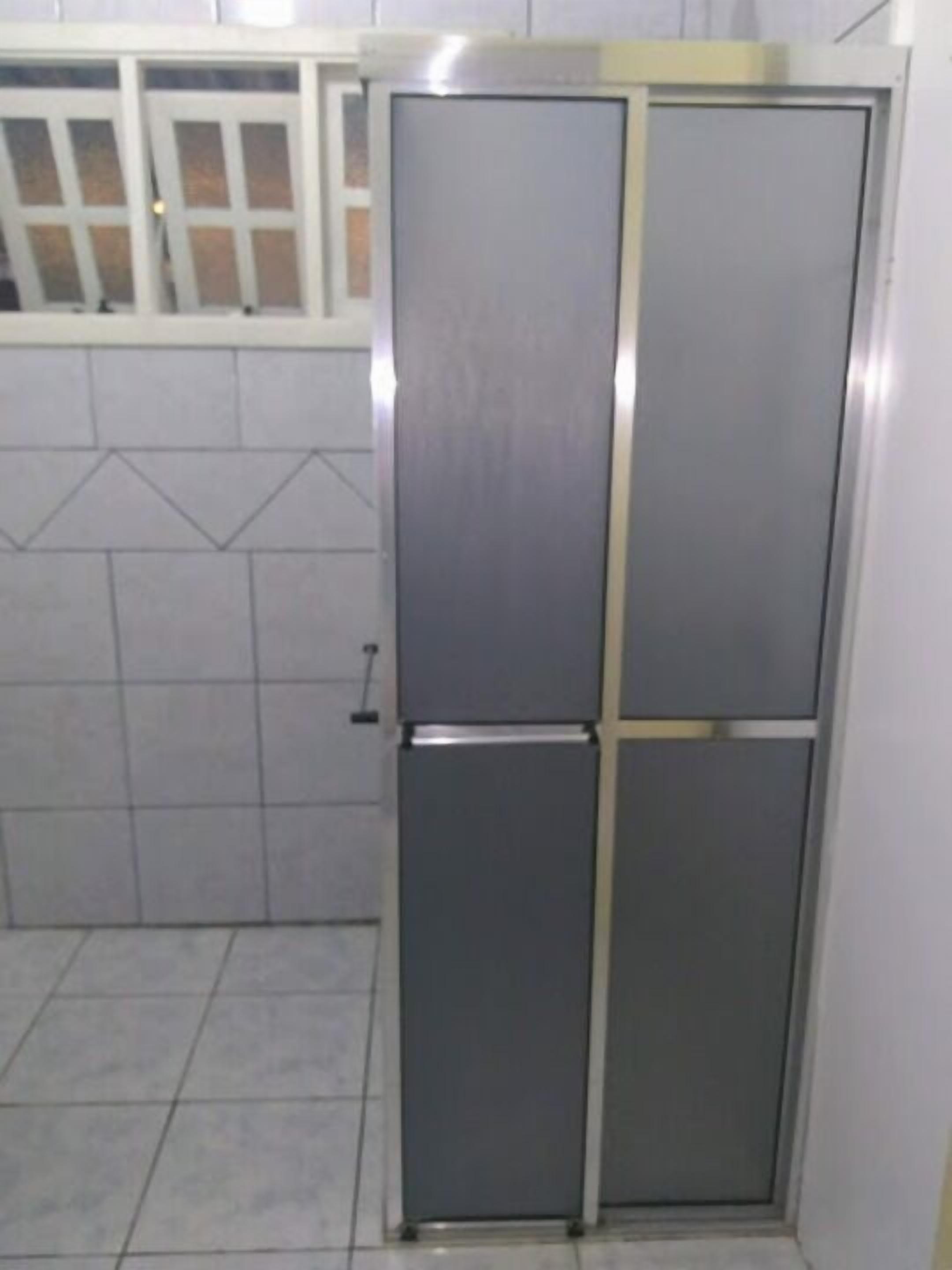 Uma geladeira compacta, com estrutura metálica e porta deslizante, localizada em uma cozinha com piso em azulejos e uma janela acima.