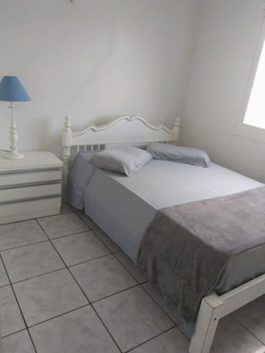 Un dormitorio con cama blanca, ropa de cama gris, una mesita de noche con lámpara y piso de baldosas.