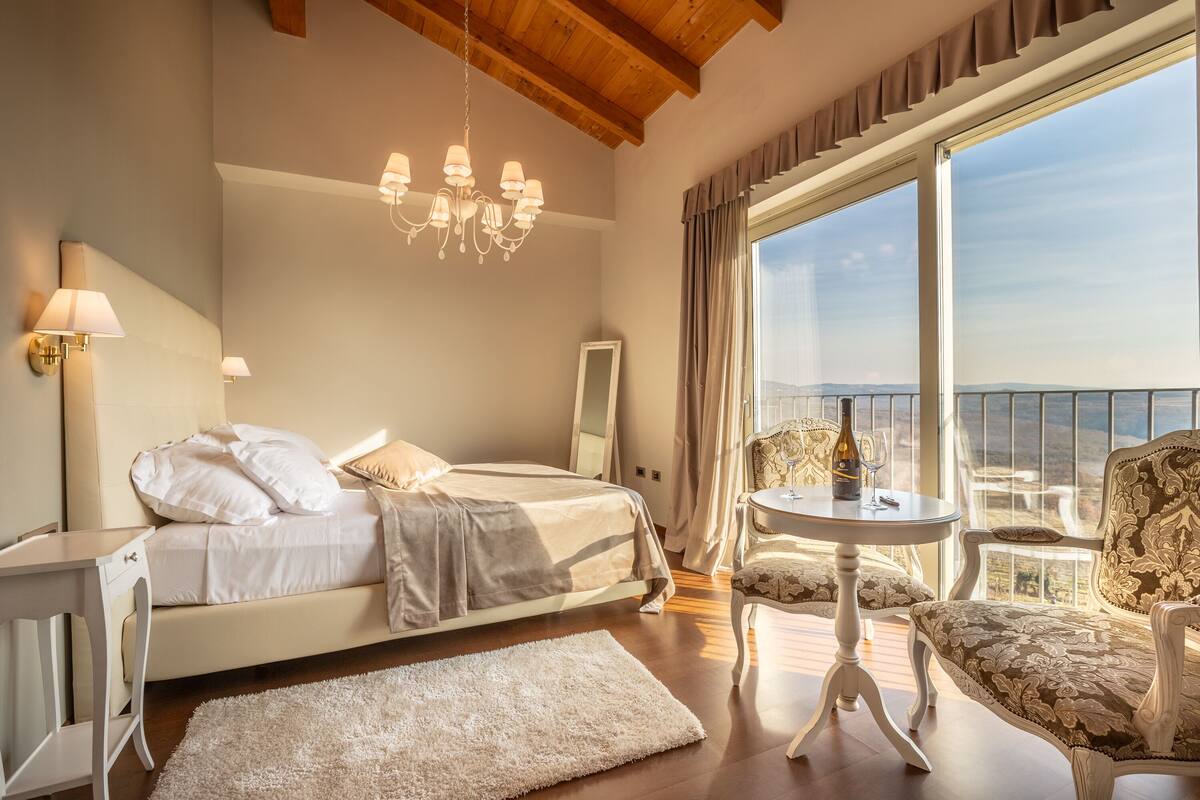 Een slaapkamer met een bed, een kroonluchter, een tafel met stoelen en uitzicht op het omliggende landschap.
