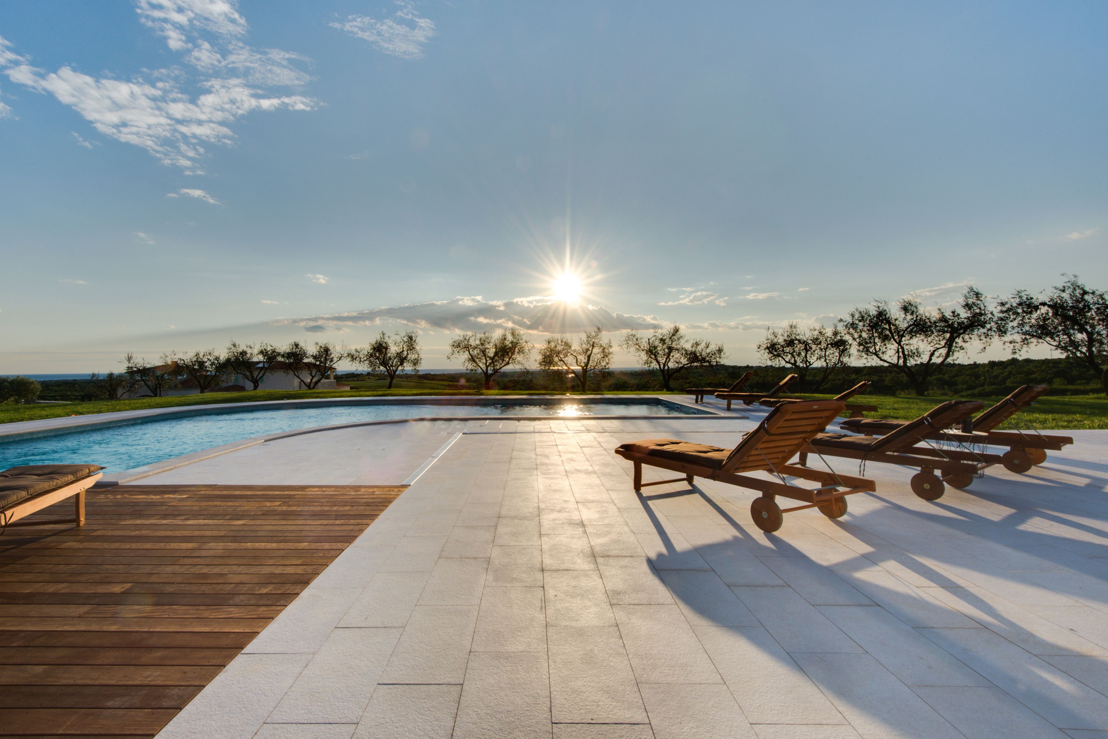 Area piscina con poltronne relax, un pontile in legno e vista sul tramonto su un paesaggio.