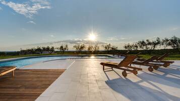 Area piscina con poltronne relax, un pontile in legno e vista sul tramonto su un paesaggio.