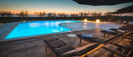 Piscina con sillones de descanso y sombrillas, rodeada de árboles y una vista al atardecer.