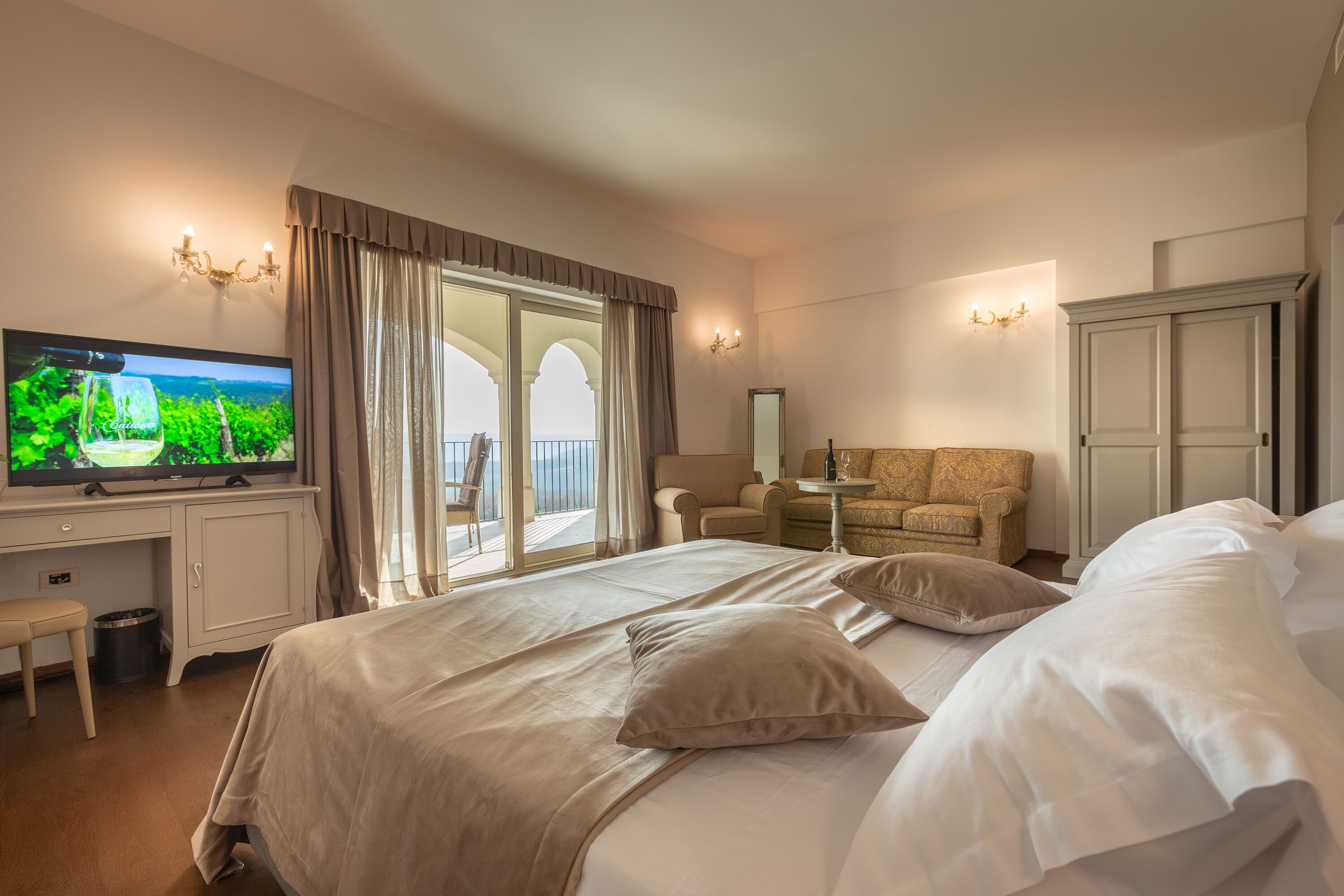 Ein Hotelzimmer mit einem großen Bett, einem Fernseher auf einem Schreibtisch, einem Sofa und einem Balkon mit Blick.