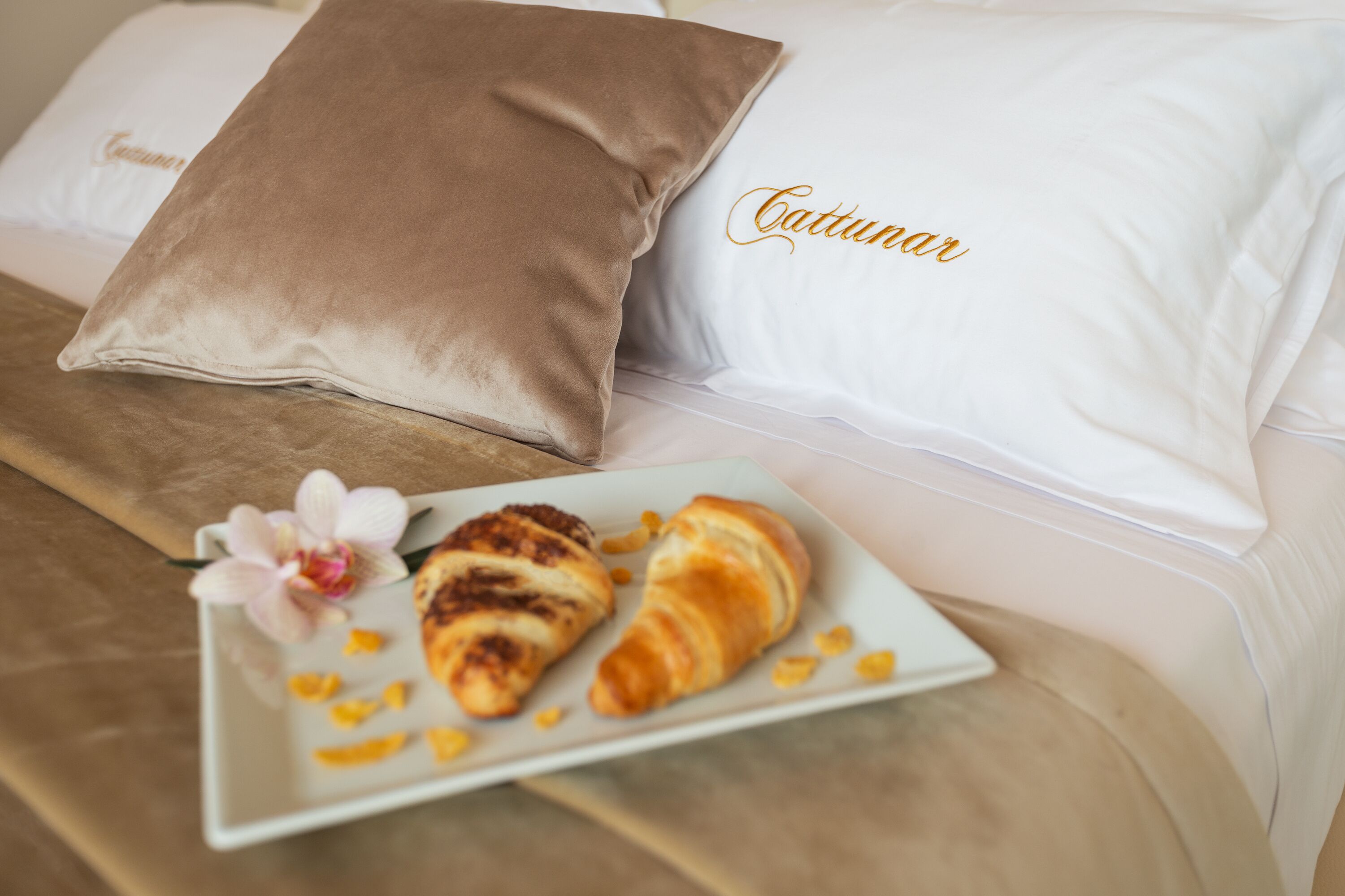 A bed with two pillows, one with the name "Cattunar" embroidered on it, and a plate with two croissants and a flower.