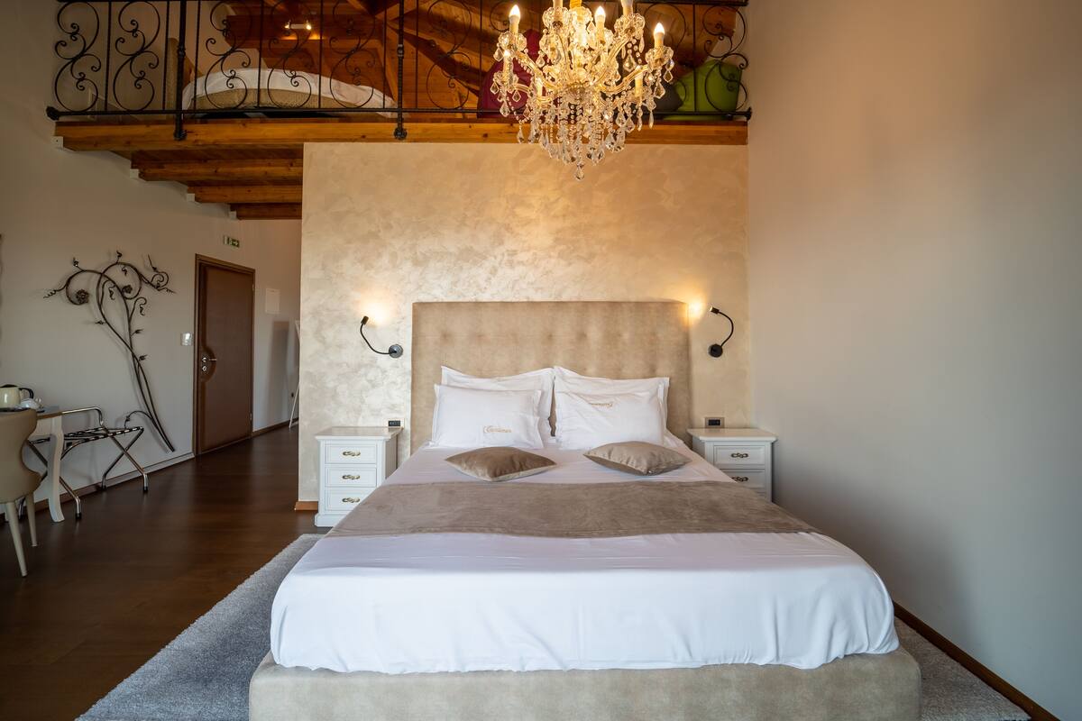 Een slaapkamer met een groot bed, houten balkenplafond, een kroonluchter en een nachtkastje met lamp.