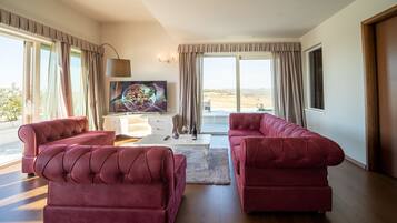 A spacious living room with two red sofas, a white coffee table, and a large TV on a white cabinet. The room has large windows with curtains and a view of the outdoors.