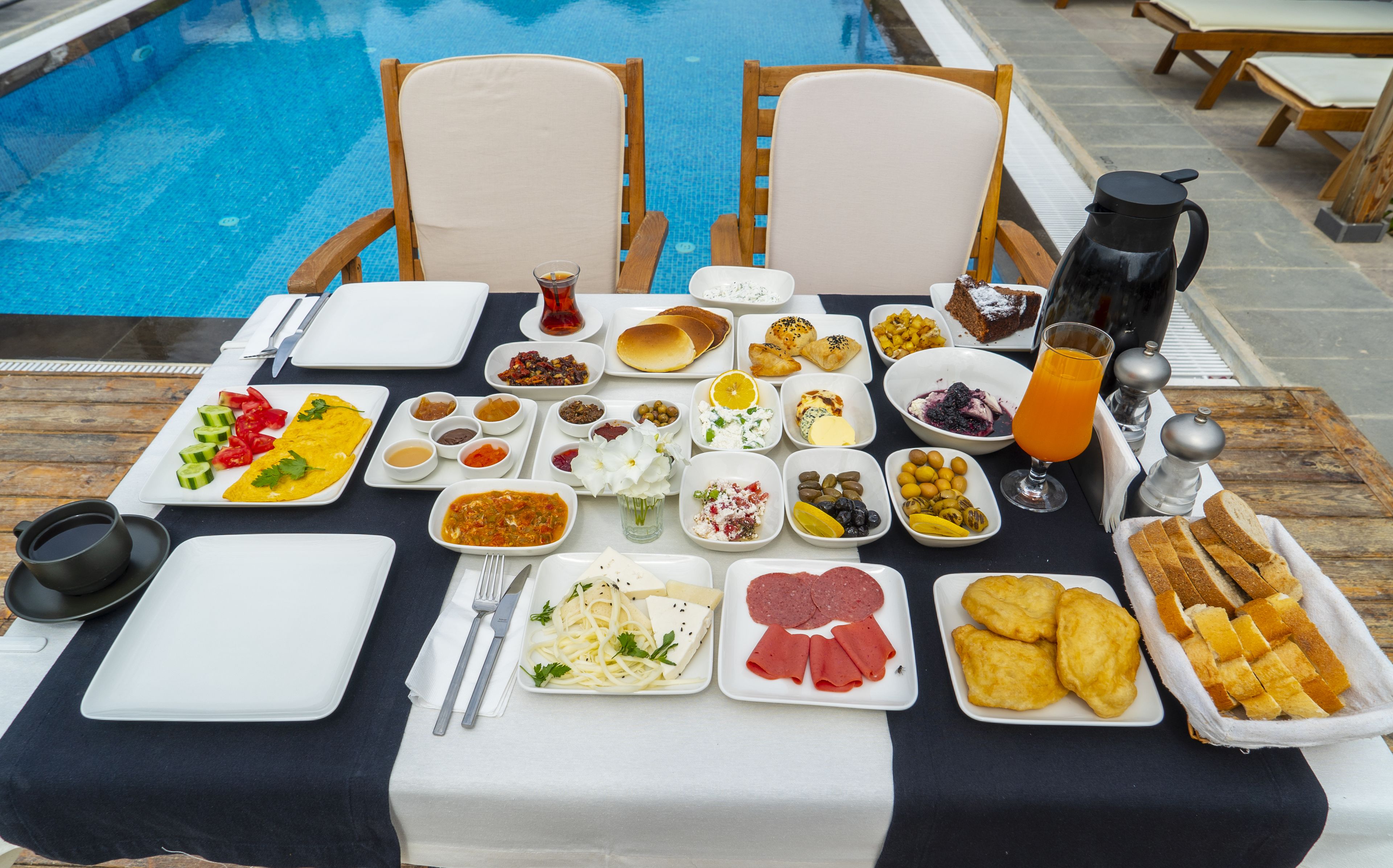 Una mesa de comedor con diversos platos y una cafetera, ubicada cerca de una piscina.