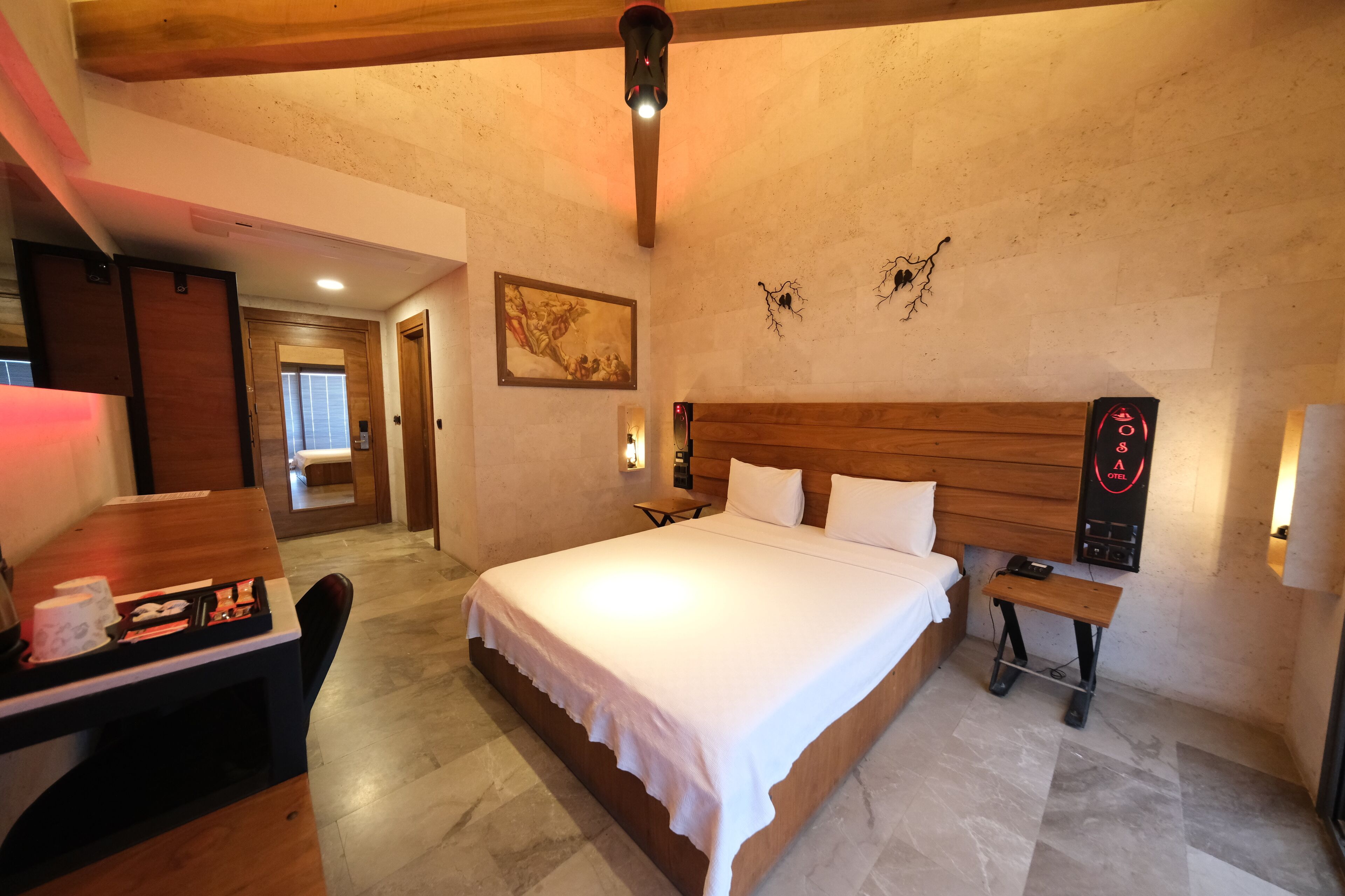A hotel room with a large bed, wooden headboard, bedside table, and a desk with a chair.