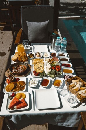 Una mesa servida con diversas opciones de desayuno, como frutas, pan y bebidas, cerca de una piscina.