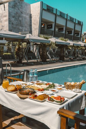 Zona de comedor junto a la piscina con una mesa dispuesta para el desayuno, con vistas a una piscina y sillones de descanso.