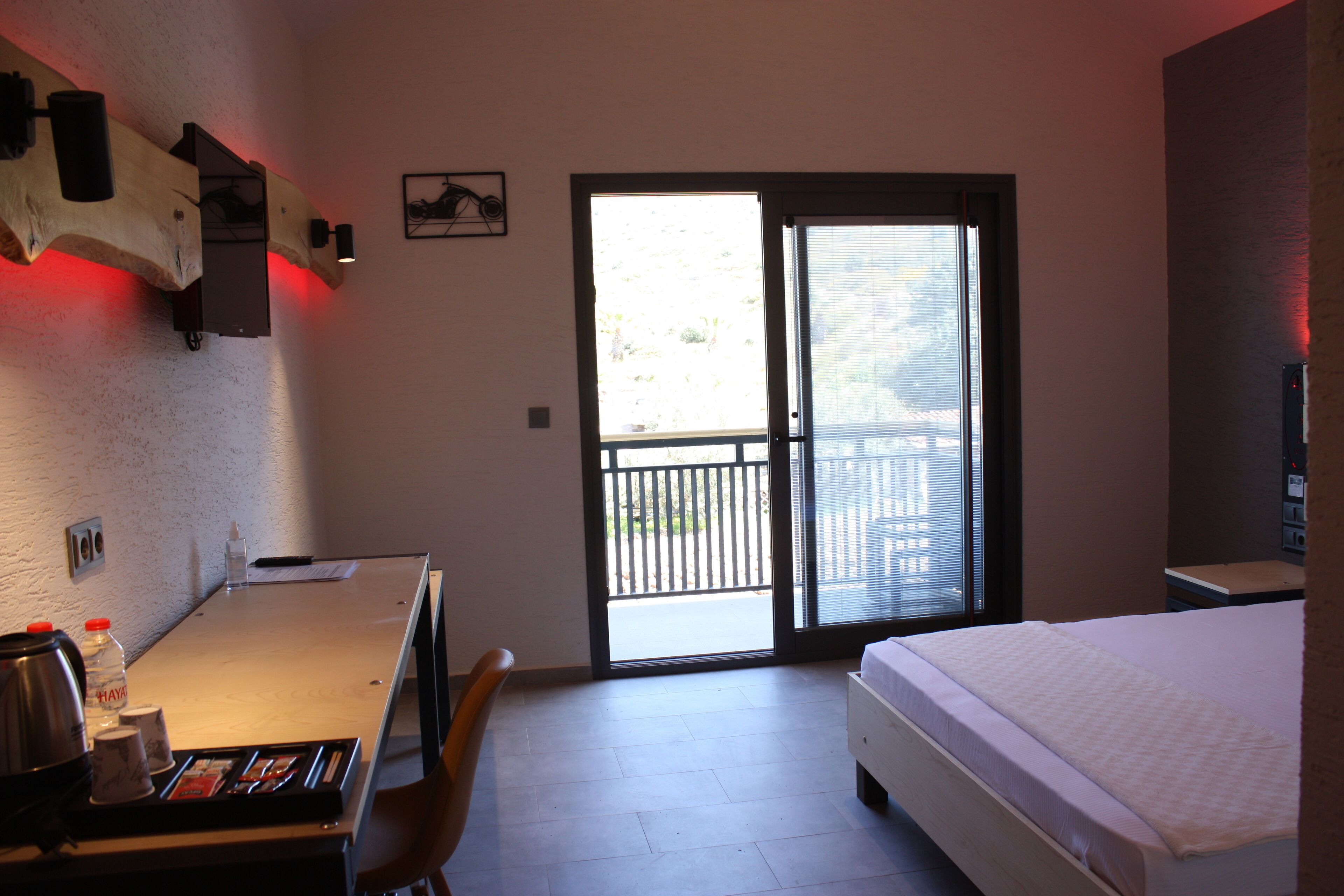 A hotel room with a bed, a desk with a chair, a coffee maker, and a bottle of water. There is a sliding door leading to an outdoor balcony.