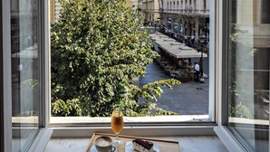 Una ventana con vista a un árbol, una mesa con un pastel y un vaso de bebida, y un edificio de arquitectura clásica.