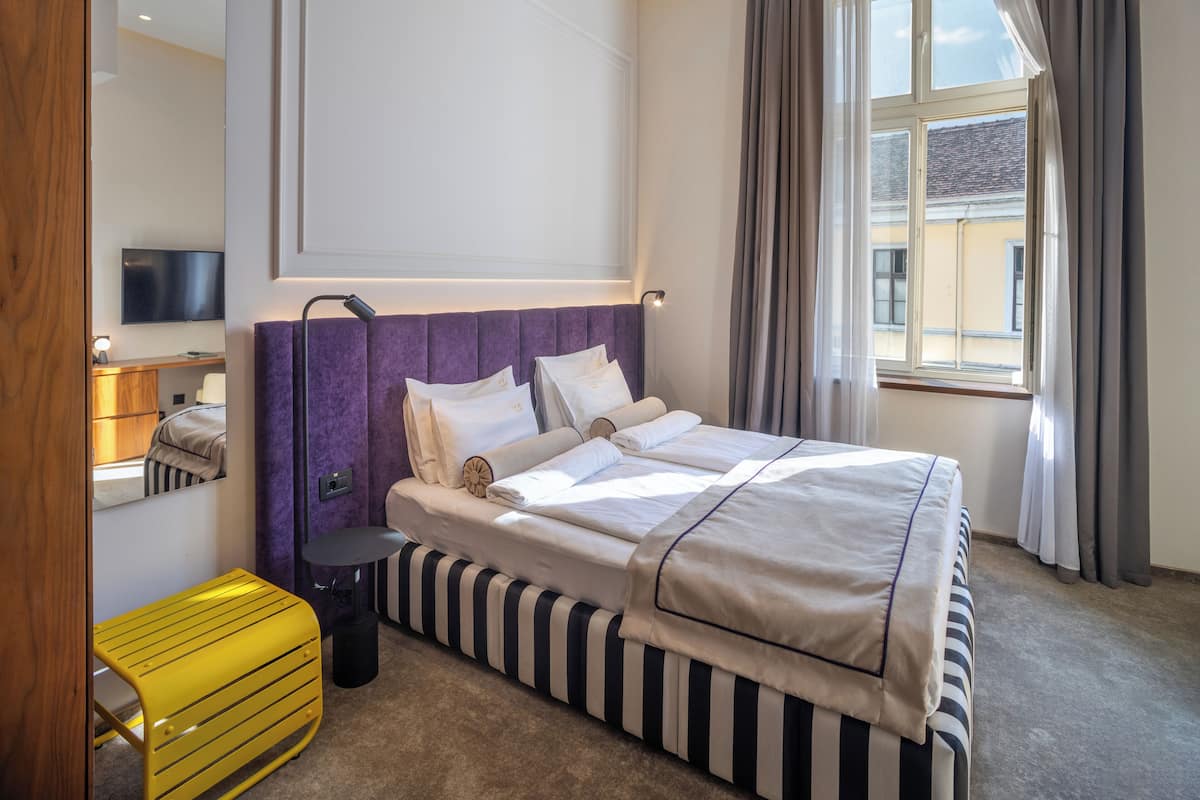 Ein Hotelzimmer mit einem großen Bett, einem Nachttisch, einer Lampe, einem Spiegel, einem Fenster mit Vorhängen und Blick nach draußen.