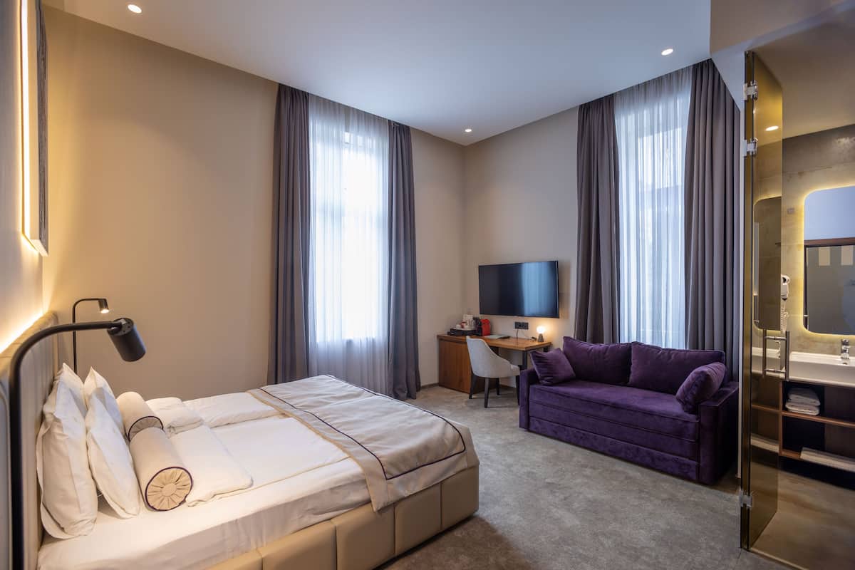 Ein Hotelzimmer mit Bett, Schreibtisch mit Fernseher, violettem Sofa und Badezimmer mit Glasabtrennung.