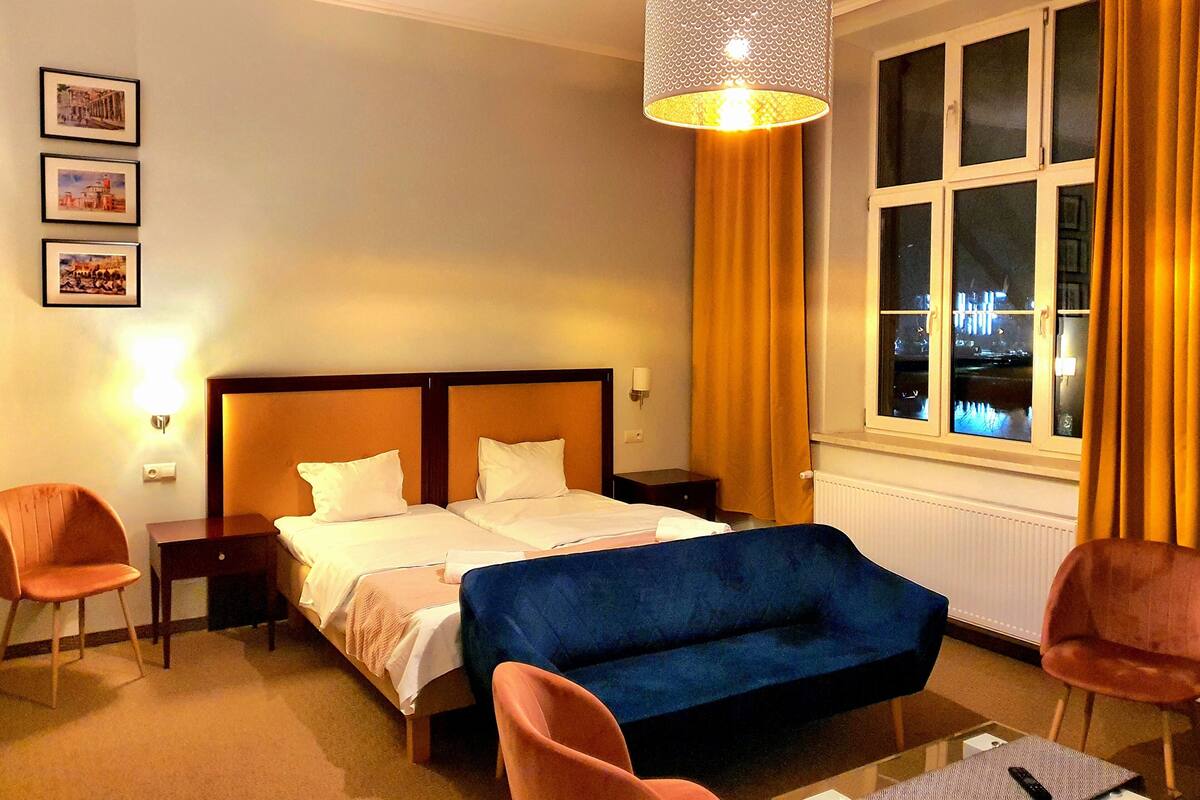 A hotel room with a bed, a blue sofa, two chairs, a nightstand, a lamp, and a window with curtains.