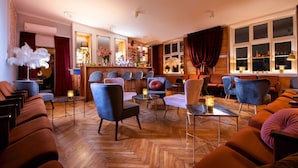 Un vaste espace salon avec de nombreux coins salon, comprenant des canapés et des fauteuils, disposés autour d’une table basse centrale. Un bar est aménagé, avec un miroir et des éléments décoratifs. La pièce est dotée d’un parquet et de grandes fenêtres avec des rideaux.