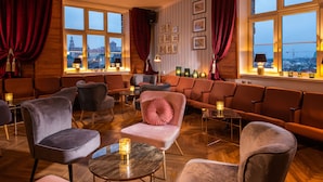 Un hall d’hôtel doté d’un espace salon avec des fauteuils et des tables confortables, de grandes fenêtres avec des rideaux et une vue sur la ville.