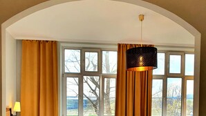 Une chambre d’hôtel avec un lit, un canapé bleu, une petite table et une chaise. De grandes fenêtres sont présentes, avec des rideaux, un radiateur et un luminaire suspendu.