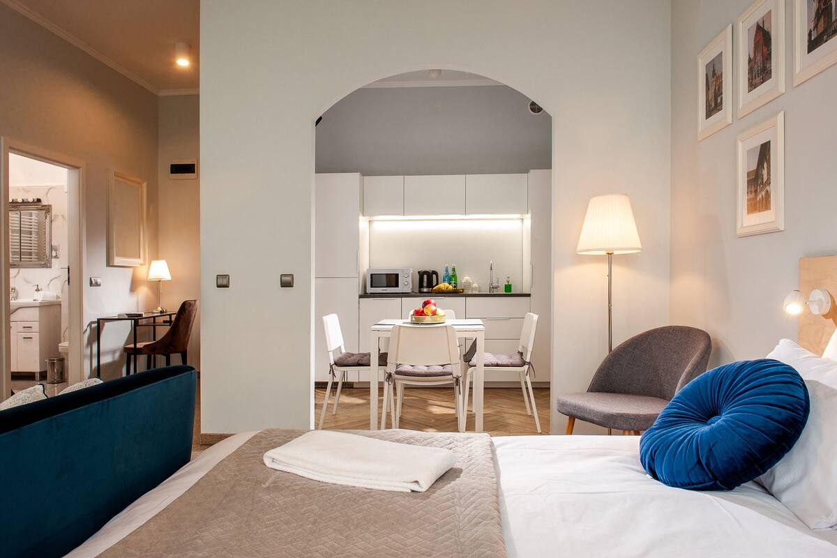 A hotel room with a bed, a desk, a chair, a lamp, and a view of the kitchen.