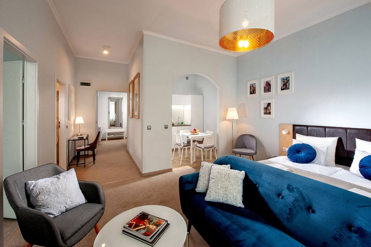 A hotel room with a bed, two armchairs, a small table, a dining area, and a bathroom.