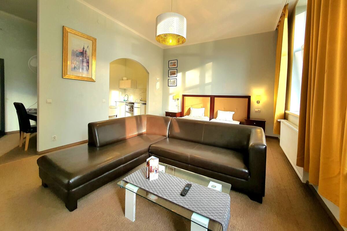 A hotel room with a brown leather sofa, a glass coffee table, a bed with white linens, a painting on the wall, and a view of the kitchen through an archway.