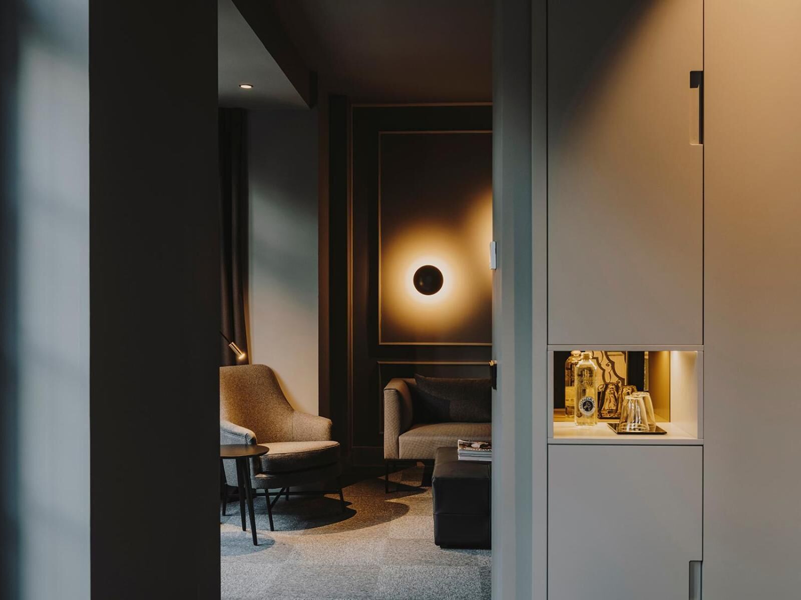 Una habitación moderna y con poca luz, con un sofá, un sillón y una mesita. Hay una lámpara circular en la pared y una estantería con objetos decorativos.