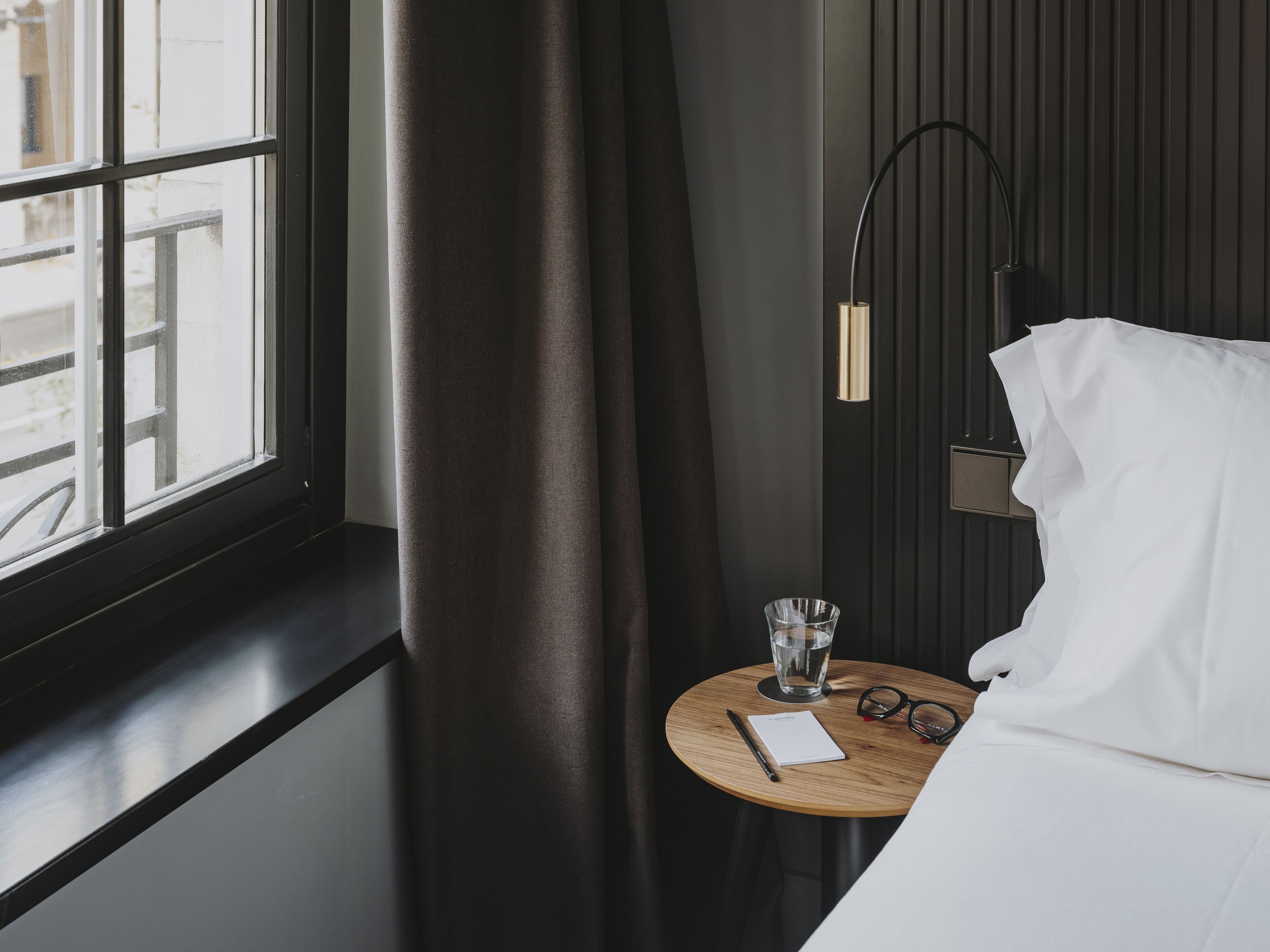 A hotel room with a bed, a bedside table with a glass of water, a pair of glasses, and a notepad.