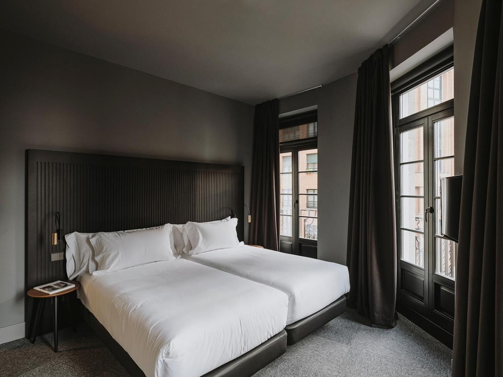 A double bed with white linens, a dark headboard, a bedside table with a lamp, and a window with curtains.